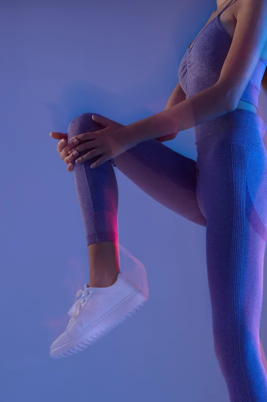 Woman sport fitness wearing textured purple activewear and white sneakers, stretching her leg against a blue and pink neon-lit background, demonstrating flexibility and health.