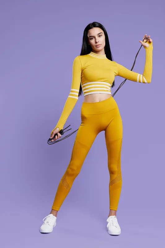 Young woman sport fitness wearing vibrant yellow activewear holding a jump rope, standing confidently against a solid purple background with white sneakers.