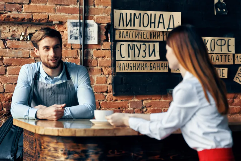 Шеф-повар в кафе или баре у кирпичной стены, посетители смотрят меню, в интерьере комнаты стоит чашка с напитком