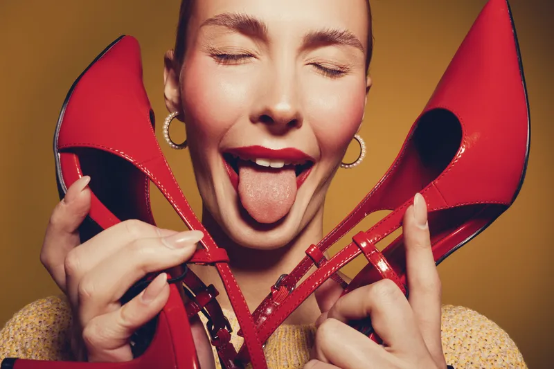 Playful woman with closed eyes holds red high heels near face, sticking out tongue. Studio portrait with warm lighting and joyful mood, concept of fashion and fun.