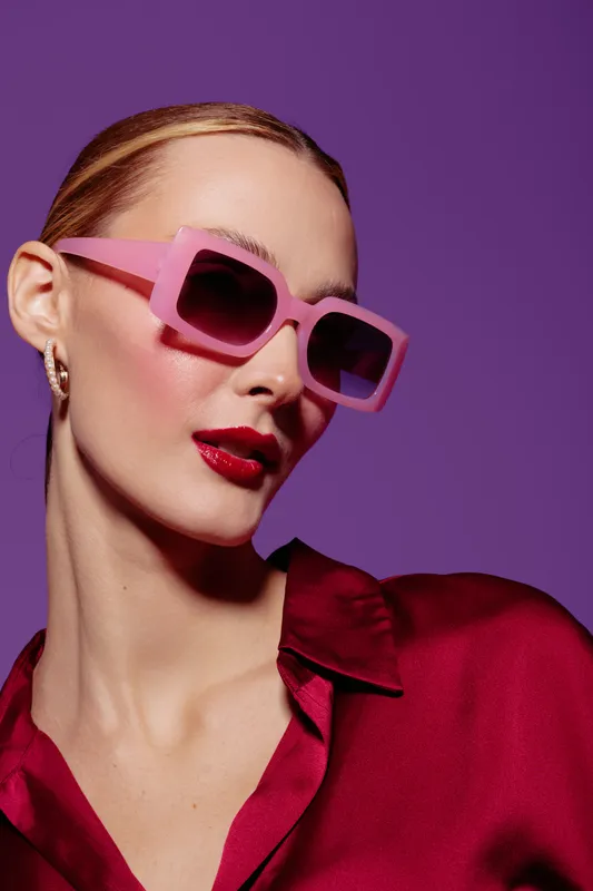 Stylish woman with pink sunglasses and red lipstick poses against purple background. Fashion concept with bold makeup, trendy accessories, and confident attitude in studio lighting.