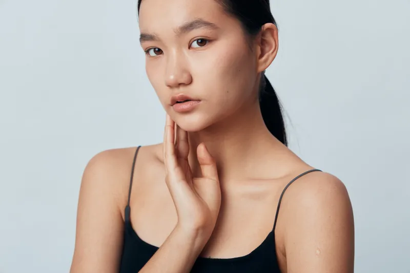 Portrait of young woman with clear skin in black top posing with hand near face on gray background. Beauty, skincare, natural look and thoughtful expression in studio lighting.