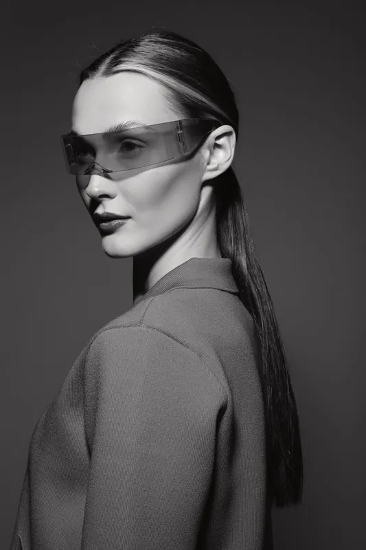 Slavic woman with pale skin and long dark hair wears futuristic transparent sunglasses and textured jacket. Studio portrait in monochrome with high contrast lighting for fashion concept.