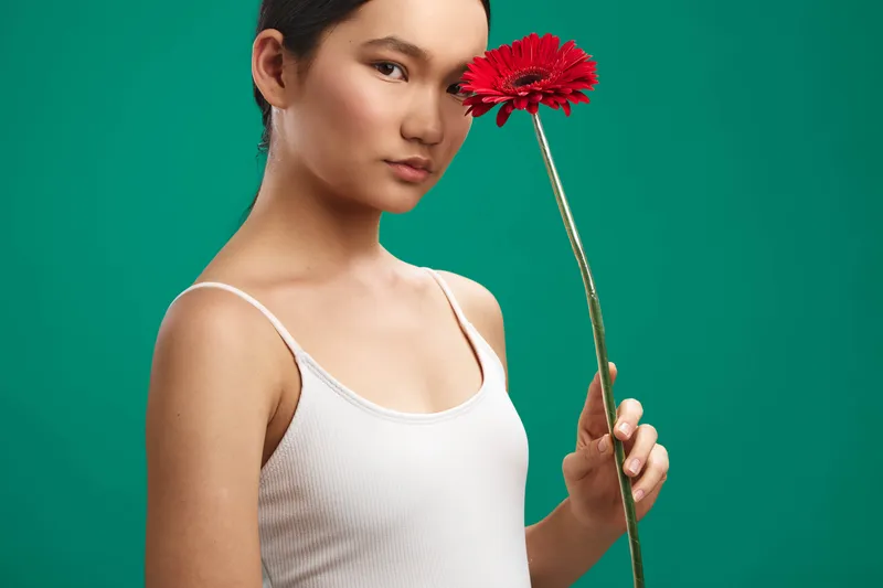 asian woman holds a bright red gerbera flower against a vivid green backdrop, creating a calm, stylish portrait with soft lighting and a gentle, composed expression.