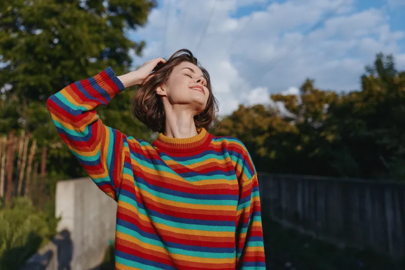 Woman in rainbow sweater outdoors, portrait in sunlight with eyes closed and hand in hair, happy and relaxed young female enjoying nature near concrete wall and trees.