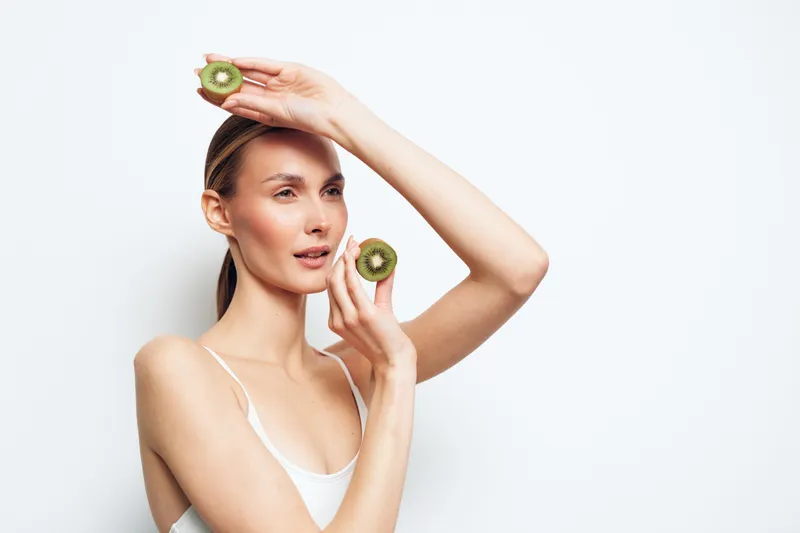 Young woman holding sliced kiwi fruit near face and head, posing against white background. Concept of healthy lifestyle, natural beauty, and skincare with fresh fruit.