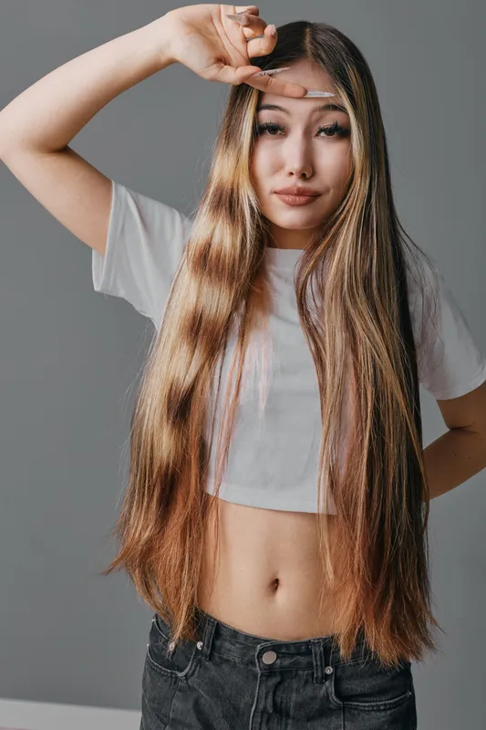 Young woman with long straight hair wearing white crop top and black jeans posing with hand near forehead, neutral background, casual fashion and expressive face portrait.
