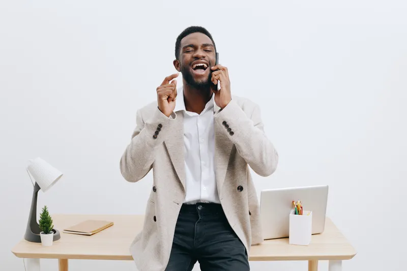 Smiling man talking on smartphone in modern office setting with white wall background, tidy desk with laptop, lamp, and plants, casual business attire, cheerful expression. People lifestyle concept, c