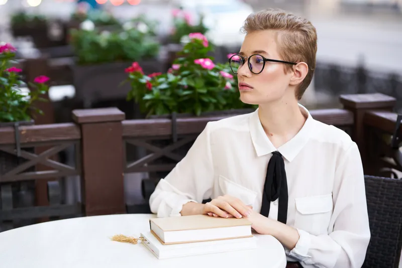 Молодая женщина в рубашке и очках с книгой за столиком в кафе