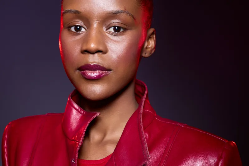 Close-up portrait of confident African woman with dark skin wearing shiny red leather jacket against dark background. Concept of fashion, beauty, empowerment, and modern style.