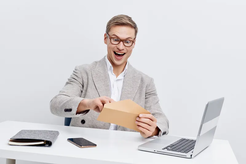 Smiling young Caucasian man in casual business attire opening an envelope during work at modern office with laptop and notebook, expressing excitement and surprise, indoor shot. People lifestyle conce