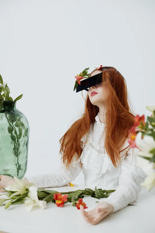 Red haired model in a white blouse with flowers, showcasing emotions of tranquility and surrealism against a minimalist background