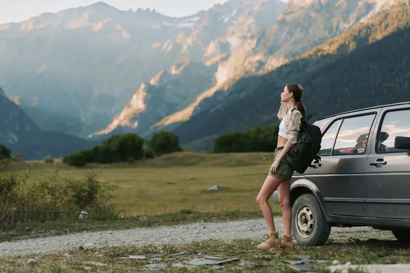 Young woman traveler with backpack stands near car on mountain road, enjoys nature view, wears shorts and boots, lifestyle adventure concept outdoors.