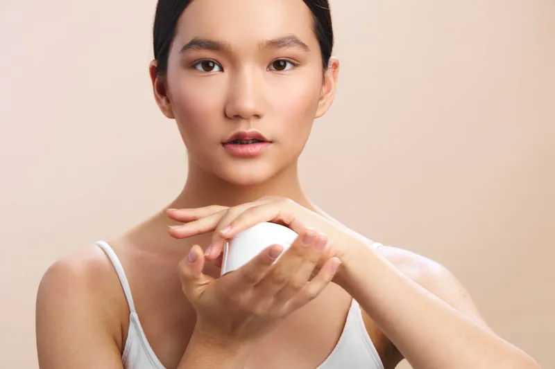 asian woman applying skincare product in a clean studio setting. She looks at the camera with a soft smile, conveying calm confidence and a gentle everyday beauty routine.