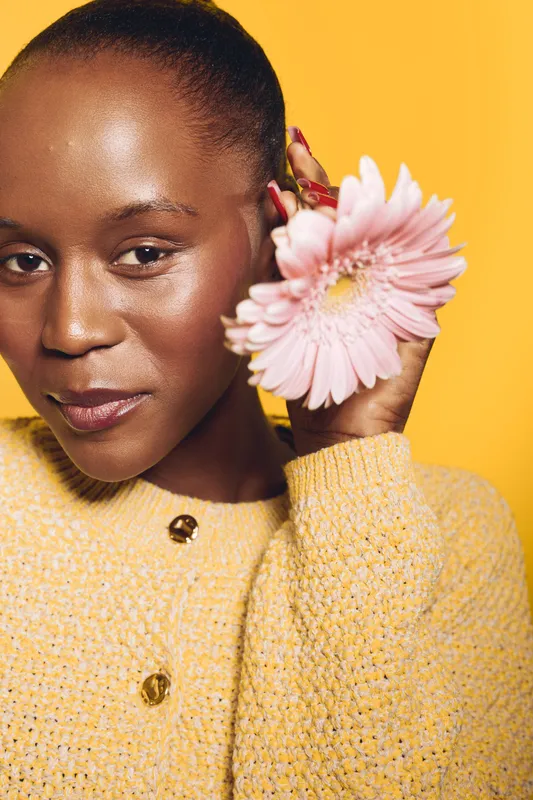 Close-up of a smiling black African woman with dark skin holding a pink flower near her face. She wears a yellow knitted sweater against a yellow background. Concept of beauty and nature.