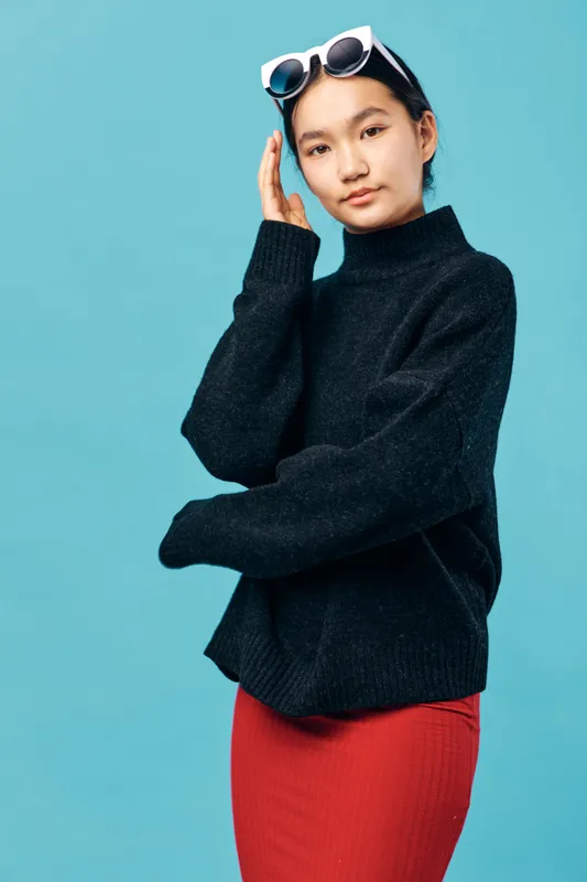 Young woman in black sweater and red skirt posing with white sunglasses on head against blue background. Fashion model with casual style and confident expression in studio lighting.