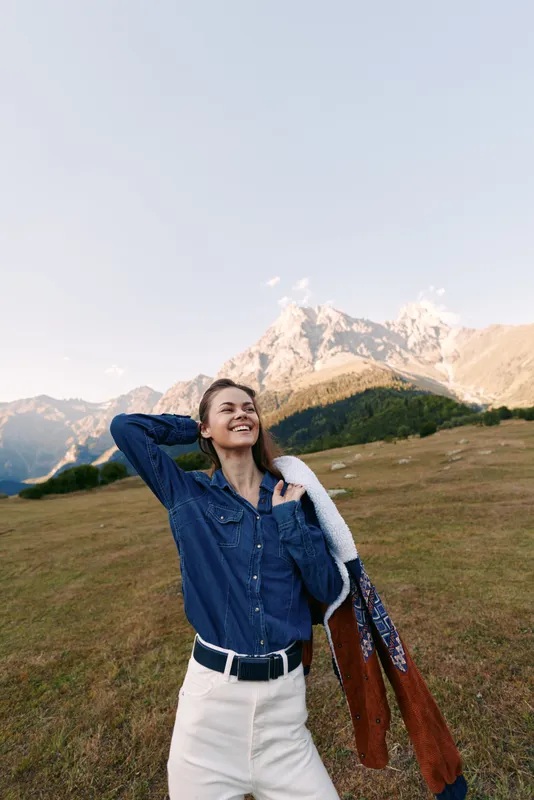 Woman in denim shirt with scarf stands smiling in meadow with mountains behind, outdoors travel portrait capturing smile, happiness, sunlight, casual style and relaxed nature moment.