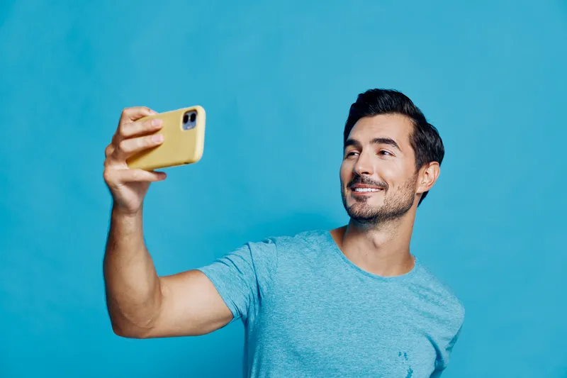 Happy cheerful young man smiling while taking a selfie with his mobile yellow phone against a vibrant blue background in a casual outfit. Lifestyle concept.
