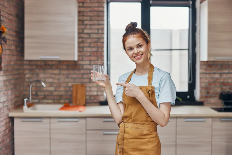 Симпатичная женщина на кухне с бокалом воды на фоне кухонной утвари и интерьера квартиры