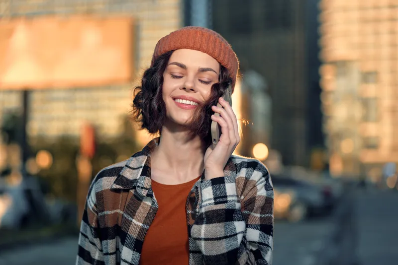 woman phone smile city beanie golden authenticity candid lifestyle emotional storytelling: young woman on mobile, relaxed and joyful in warm golden hour glow, mindful living portrait in urban street