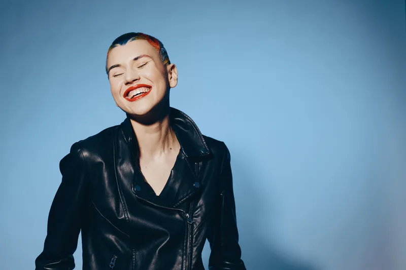 Smiling woman with short hair wearing a black leather jacket standing against a blue background, expressing joy and confidence with bright red lipstick.