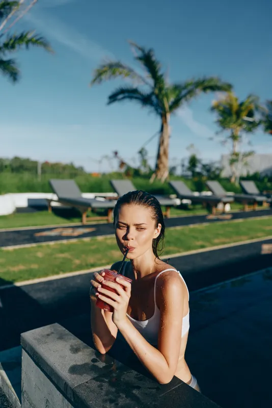 Vibrant lifestyle portrait of a woman by a pool, sipping a refreshing drink, sunny poolside moment, relaxed posture, warm breeze, bright colors, tropical backdrop