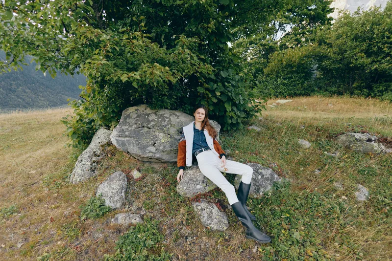 Woman reclining on mossy rocks under a leafy tree in a grassy field, wearing riding boots and a casual jacket; countryside outdoor portrait conveying a relaxed nature escape.