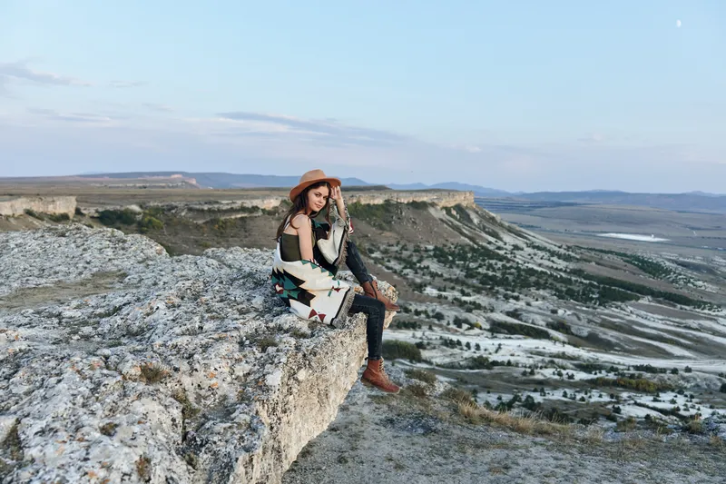 Женщина, стоящая на краю скалы, задумчиво смотрит на бескрайнюю долину внизу на фоне величественных гор
