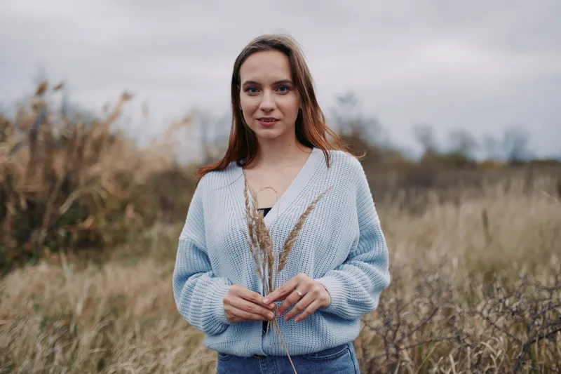 A natural portrait of a young woman standing in a windswept field, holding dried grasses and wearing a cozy sweater, conveying quiet authenticity and subtle credibility in simple rural life.