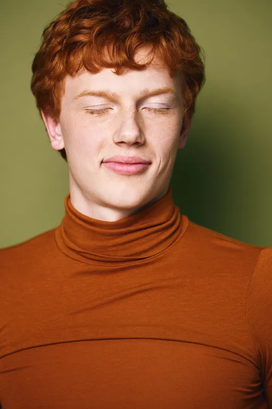Young man with red hair and freckles wearing brown turtleneck smiling with closed eyes against green background, portrait of casual confident person with relaxed expression and natural style.