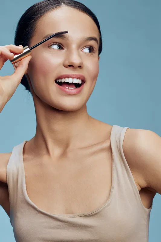 Young woman applying eyebrow makeup, showcasing beauty and confidence against a blue background, emphasizing modern personal grooming and self expression
