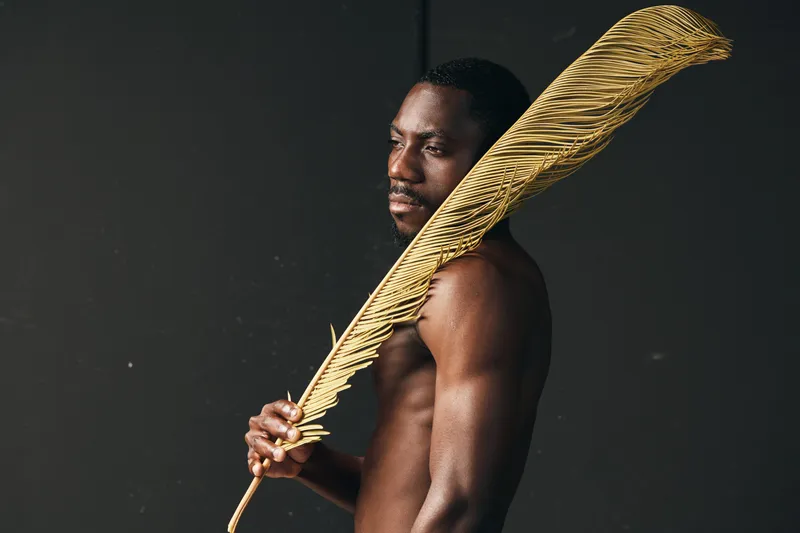 African man with dark skin holding large golden feather in creative conceptual visual style. Portrait of shirtless male model with artistic prop on dark background for fashion and art.