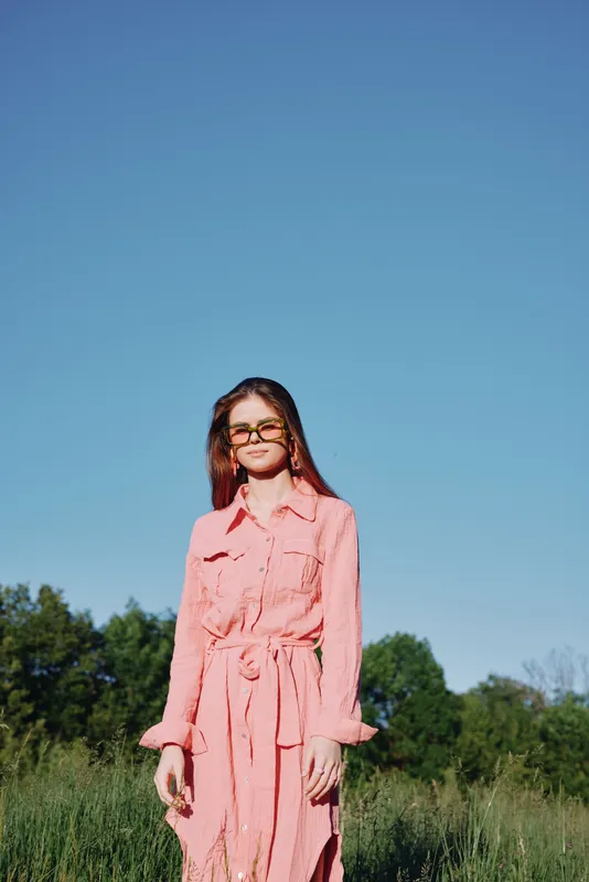 Young woman in stylish pink dress against clear blue sky, exuding confidence and joy in a lush green field, perfect for fashion and lifestyle themes