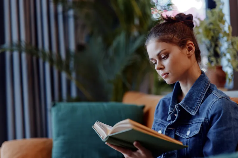 Отдыхая дома, женщина читает книгу на диване перед растением в уютной обстановке гостиной