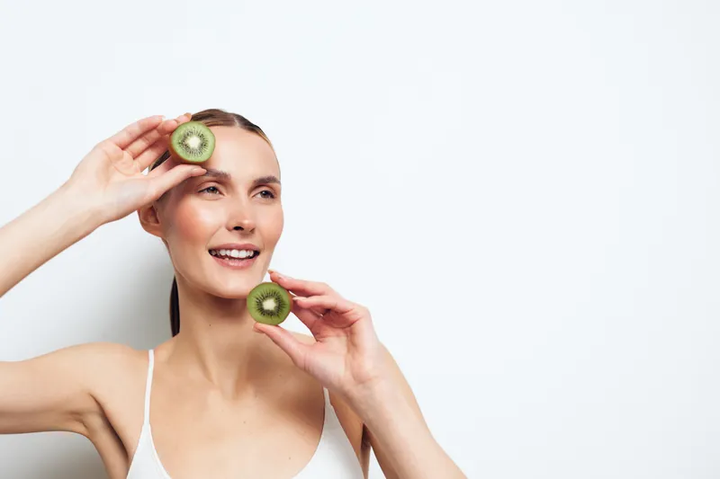 Young woman with long hair smiles joyfully holding two halves of a kiwi fruit near her face against a white background in a fresh healthy lifestyle concept.