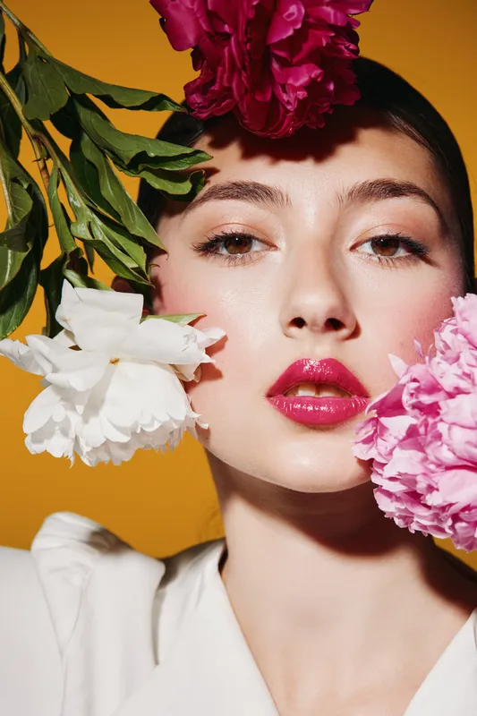 Close-up of a young woman with glossy red lips posing among pink and white flowers against an orange background, evoking themes of beauty creativity and fashion glamor.