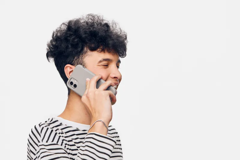 smiling man talking on smartphone wearing striped shirt with curly hair and light background, expressing happiness and casual communication.