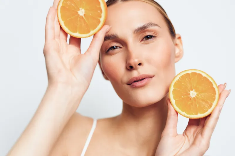 Close up of a woman holding orange halves near her face, bright studio lighting, fresh citrus tones, clear skin, minimal white background, cheerful and natural mood