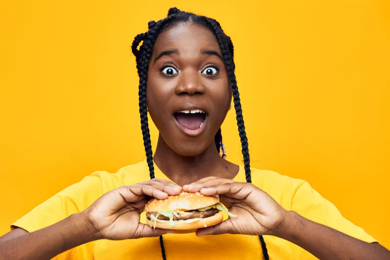 Excited girl with braids holding a delicious hamburger against a vibrant yellow background, showcasing joyful emotions and a fun food concept