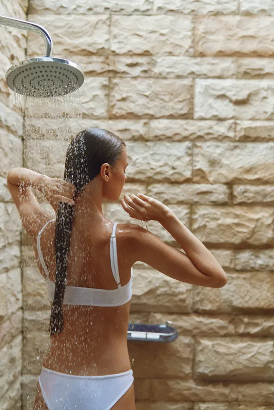 woman taking shower with water falling on her back, long dark hair, wearing white underwear, natural stone wall background, morning light