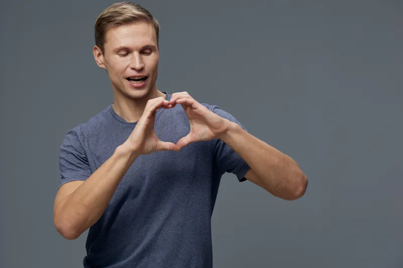 Smiling man making heart gesture casual wear blue shirt studio gray background mental health and gadgets concept