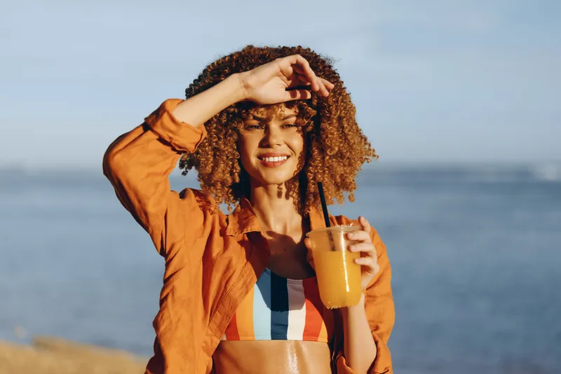Smiling woman with curly hair on beach sand in summer sunlight, holding a drink of juice in a cup, relaxed posture, seaside ocean horizon behind, holiday relaxation mood, warm sunny day outdoors