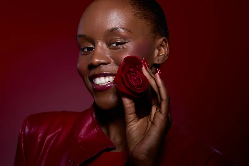 Close-up portrait of a smiling black woman with dark skin holding a red rose near her face. She wears a red leather jacket and lipstick, against a deep red background, concept of beauty and elegance.