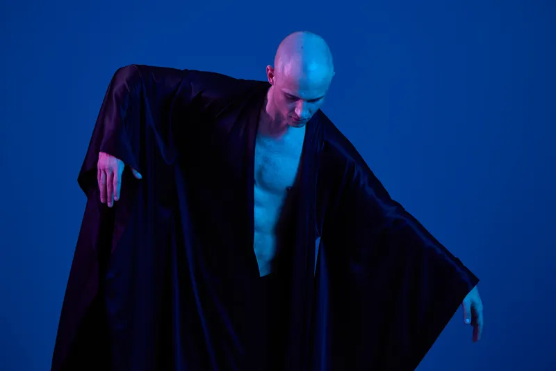 A man in a flowing black garment poses dramatically against a blue background, showcasing elegance and movement in a striking black and white artistic style.
