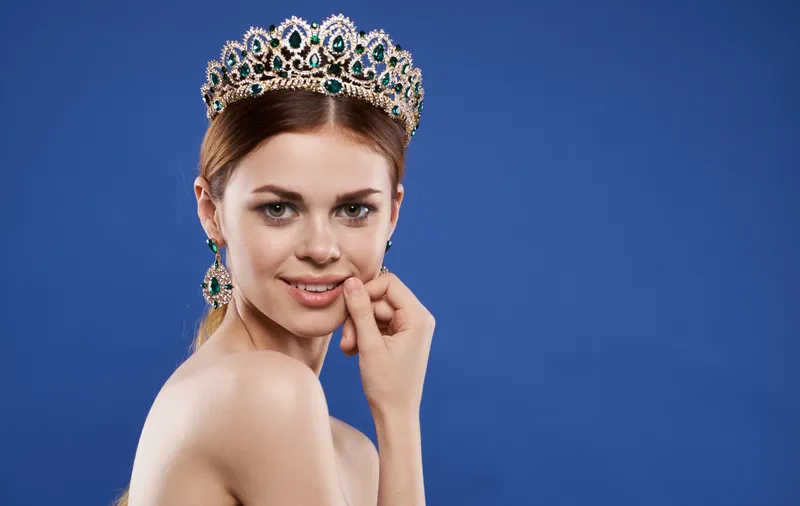 Портрет женщины в короне на голове, модель с макияжем, делающая жест рукой на синем фоне