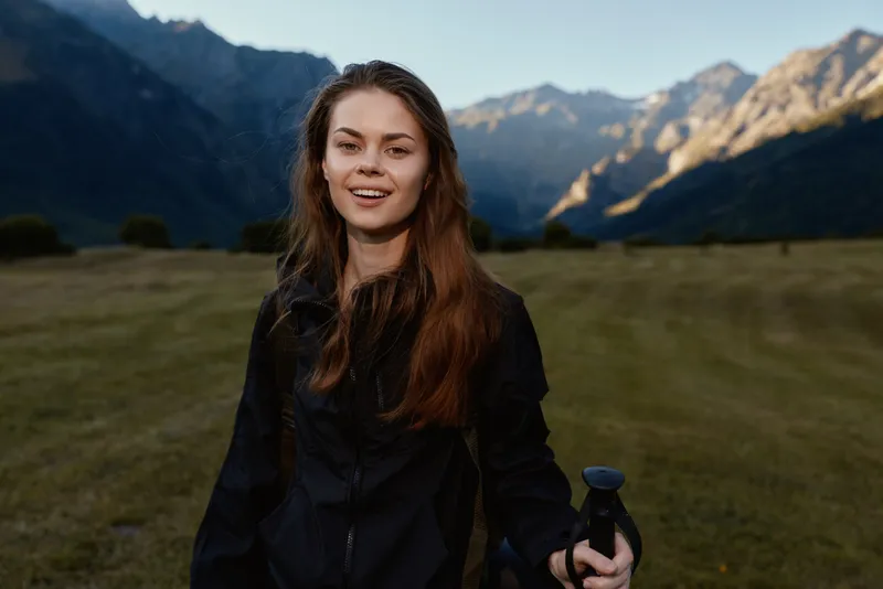 A young woman stands in a mountain meadow, holding a bottle and smiling at the camera. The scenic alpine backdrop creates a calm, adventurous atmosphere.