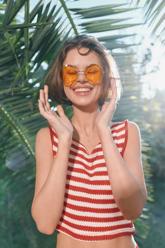 A cheerful woman wearing a striped red and white top and round orange sunglasses smiles joyfully in sunny outdoor surroundings, exuding carefree summer vibes and relaxed fashion.