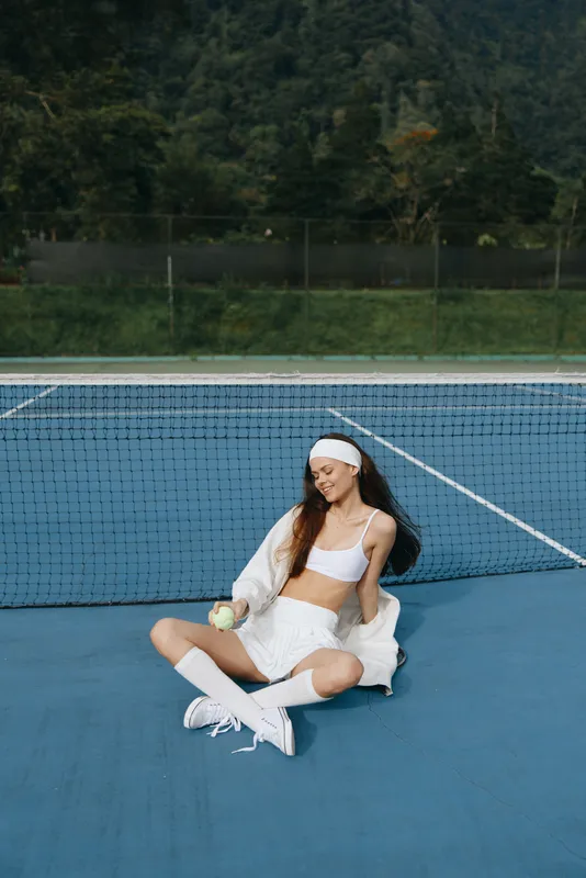 Young woman in sportswear sitting on a tennis court, smiling with a tennis ball in hand, sports lifestyle, vibrant colors, seasonal fun, healthy lifestyle