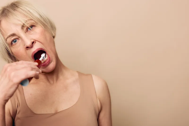 Blonde woman in beige tank top brushes teeth with white toothbrush, showing dental care routine on neutral background. Concept of oral hygiene, self-care, and healthy lifestyle for adults.