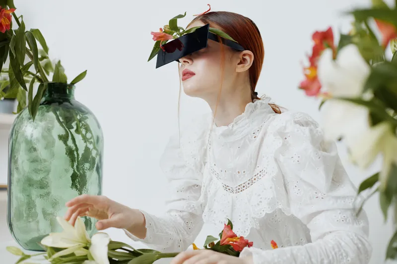 A woman in a white lace dress arranging flowers, wearing stylish glasses with a floral design, surrounded by greenery and white blooms, evoking a sense of creativity and nature